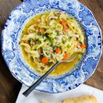 A blue and white bowl of chicken noodle soup on a wooden table with torn baguette near by.