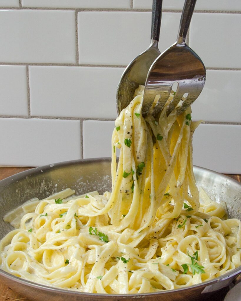 Servers lifting fettuccine alfredo out of a stainless steel skillet.