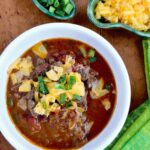 Chunky Beef Chili in a white bowl with scallions and Cheddar cheese near by.