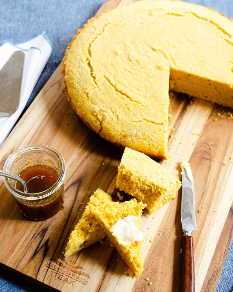 Cornbread with wedges cut out of it on a wooden cutting board with honey on the side.