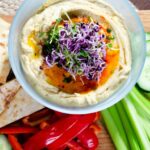 Hummus in a bowl with sprouts on top and vegetables and pita around the side.