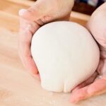 A pizza dough ball being shaped by two hands on a wooden countertop.