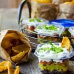 Individual 7-layer dips on a wooden table with bags of corn chips near by.