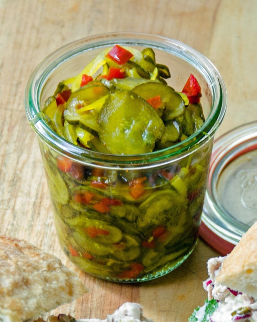 A glass jar of bread and butter pickles, lid open on a wooden table.