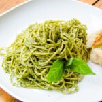 Spaghetti with basil pesto on a white plate with bread and two leaves of basil.