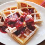 Waffles with fruit compote on top, on a white plate with an orange napkin below.