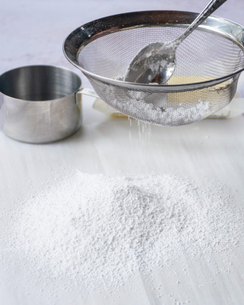 A spoon pushing powdered sugar through a strainer onto a piece of parchment paper.