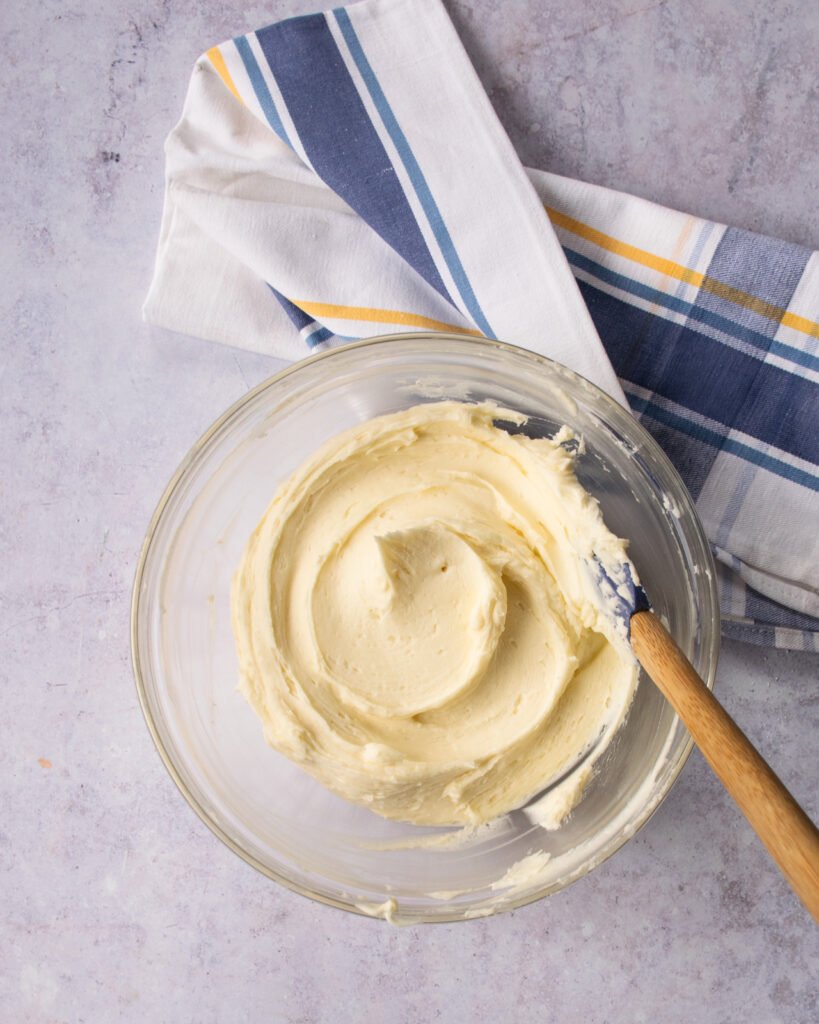 Looking down on a bowl of buttercream frosting with a spatula sticking out of the bowl and a blue and yellow towel next to it.