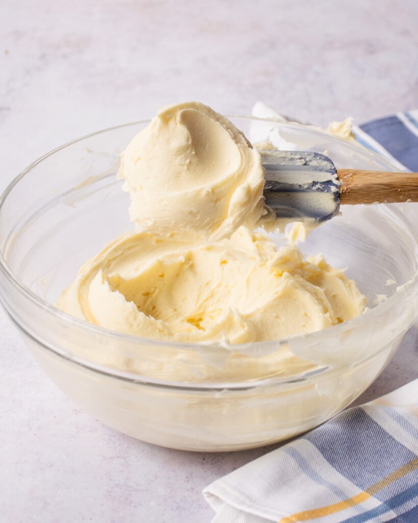 A blue spatula lifting buttercream frosting out of a bowl.