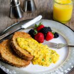 Scrambled Eggs on a piece of toast with berries, orange juice and a salt and pepper shaker on the side.