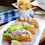 Three Lobster Rolls on a white plate with potato chips in the background.