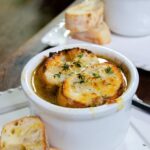 French Onion Soup in a small white cup with bread on the side.