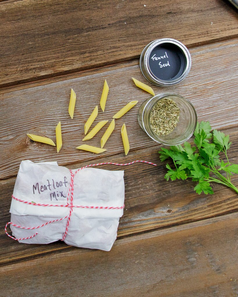 A packet of meatloaf mix, some dried penne, parsley and fennel seed on a wooden table.
