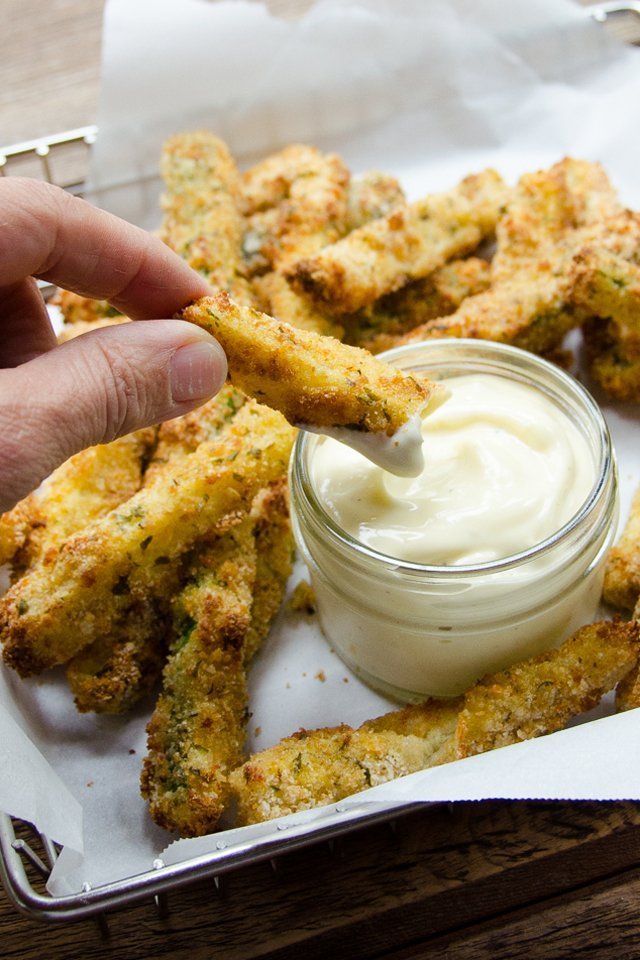 A hand holding a zucchini fry, dipping it into a jar of dip with other zucchini fries around.