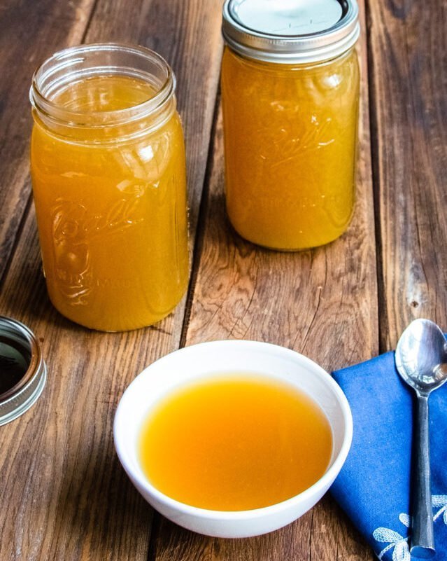 Two jars of bone broth on a wooden table with a bowl of broth in front of them.