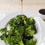 Air-fried broccoli on a white plate with soy drizzle being poured on it.