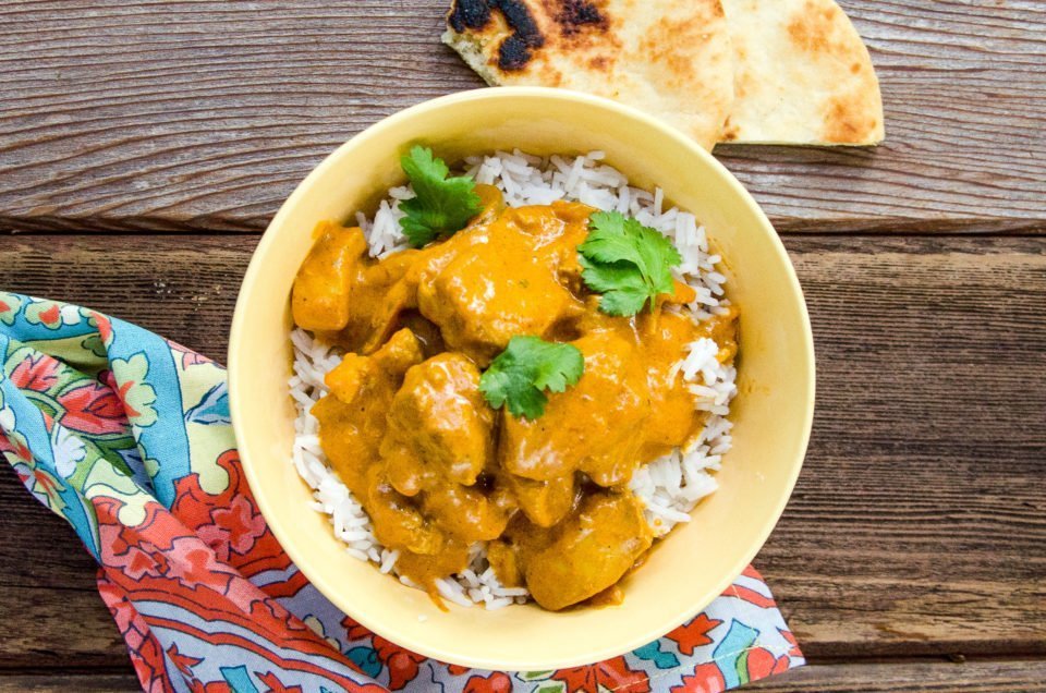 Butter chicken on rice in a yellow bowl on a wooden table, with naan on the table and a colorful napkin.