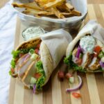 Chicken gyros on a striped wooden board with french fries in the background.