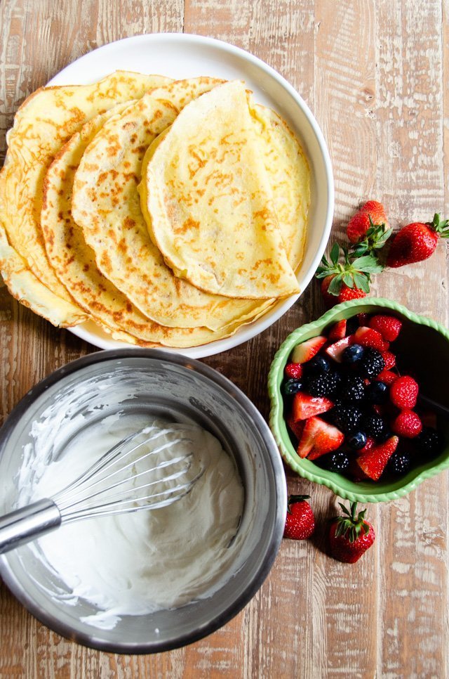 Crêpes on a white plate with a bowl of berries and a bowl of whipped cream with a whisk.