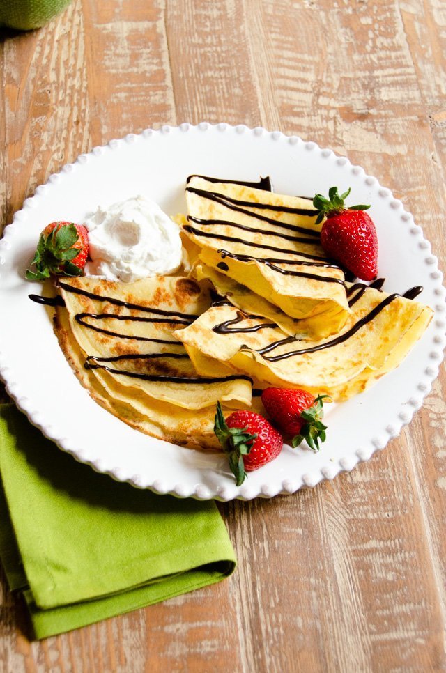 Crêpes folded in quarters on a plate with strawberries and chocolate sauce drizzled on top.