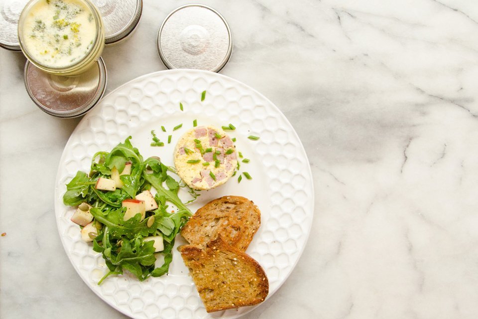Instant Egg Bite on a white plate with toast and salad greens tossed with apple. All on a marble table with jars of egg bites on the side.