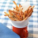 Skinny fries in a paper cone in a red cup on a blue checkered tablecloth.