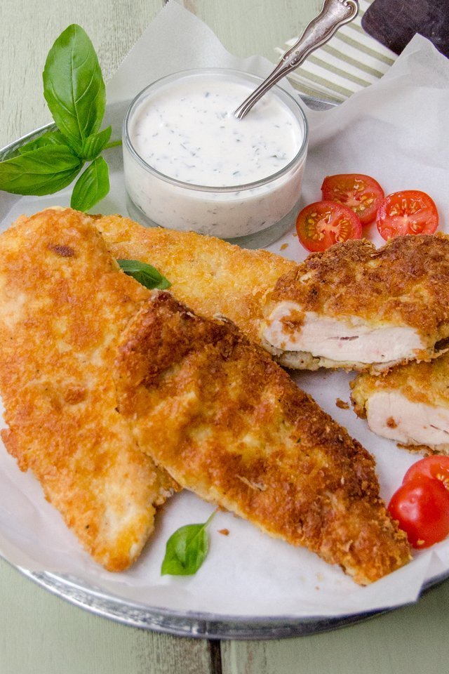 Gluten-free breaded chicken cutlets on a white plate with tomatoes and basil and a basil buttermilk dressing on the side.