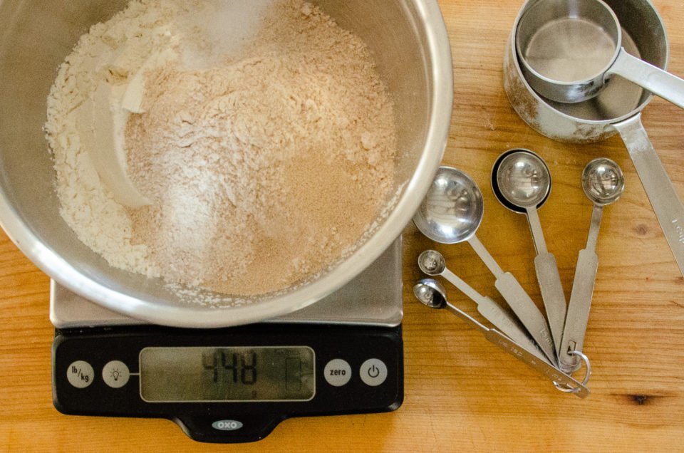 Ingredients for whole wheat no knead bread in a stainless steel bowl on a scale with measuring cups and spoons.