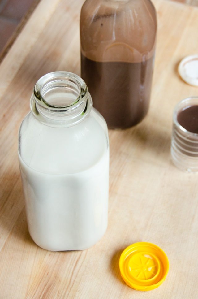 A bottle of homemade oat milk ion a table with a bottle of chocolate oat milk in the background.