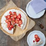 Looking down on a pistachio pavlova with strawberries on a parchment lined plate with a wedge cut out and on a smaller white plate.
