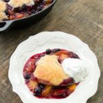 Peach and Blueberry Cobbler on a white plate with a cast iron pan filled with the rest behind it.