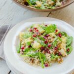 Quinoa salad with spinach, cauliflower, avocado, pecans and pomegranate seeds on a stack of white plates with a wooden bowl of salad in the background.