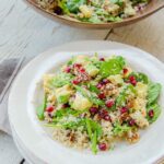 Quinoa salad with spinach, cauliflower, avocado, pecans and pomegranate seeds on a stack of white plates with a wooden bowl of salad in the background.