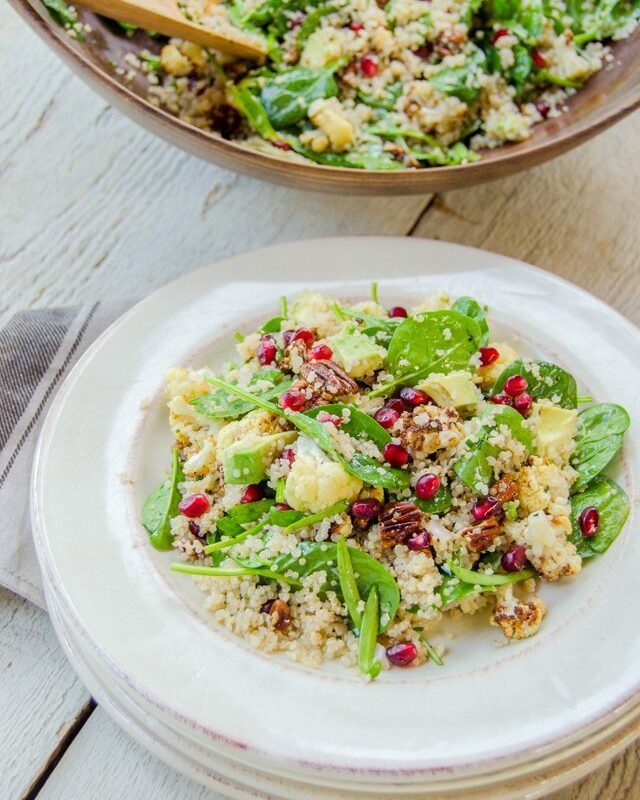 Quinoa salad with spinach, cauliflower, avocado, pecans and pomegranate seeds on a stack of white plates with a wooden bowl of salad in the background.