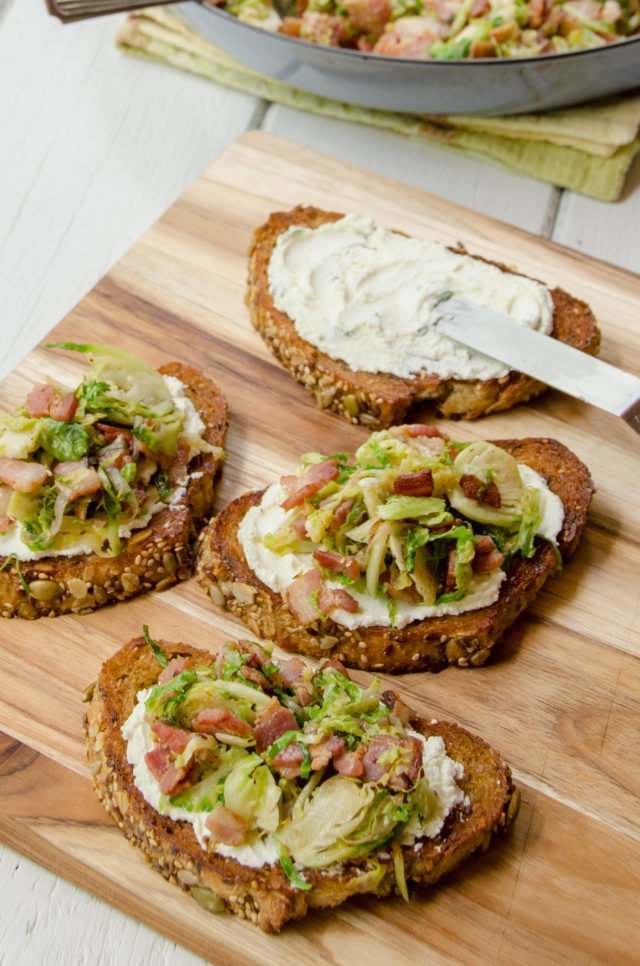 Three slices of Shaved Brussels Sprouts Toast on a wooden cutting board. A fourth toast being assembled with goat cheese spread on top.