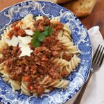 Pressure cooker bolognese sauce over rotini in a blue and white bowl with bread on the side.