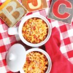 Creamy Tomato Macaroni in two small white bowls on a red and white checkered cloth with coasters "B", "J" and "C".