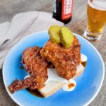 Nashville Hot Chicken on a blue plate served with white bread, cool dressing and dill pickles; a beer in the background.