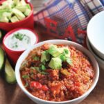 Quinoa chili in a white bowl with sour cream and avocado in dishes near by.