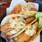 Spicy Fish Street Tacos with sriracha slaw on top in a parchment lined basket with lime wedges