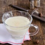 Béchamel Sauce in a glass sauce boat on a wooden table with a grater, nutmeg, salt and pepper in the background.