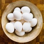 A ceramic bowl of white eggs on a wooden cutting board.