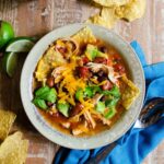 Chicken Tortilla Soup with a blue napkin, spoon, lime wedges and tortilla chips on a wooden table.