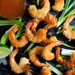 Coconut shrimp on black marble with duck sauce, scallions and lime wedges.
