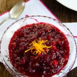 Cranberry Sauce in a glass dish with a spoon and plate on a wooden table.