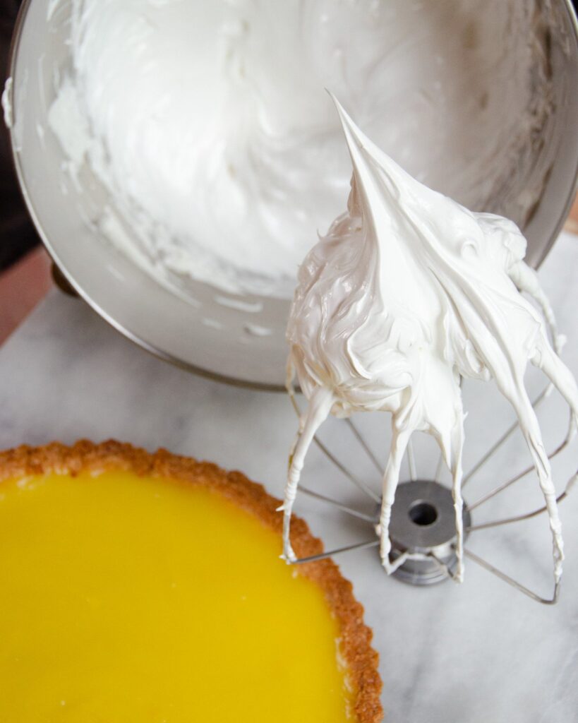Stiff peaks of meringue on a beater next to a lemon meringue tart.