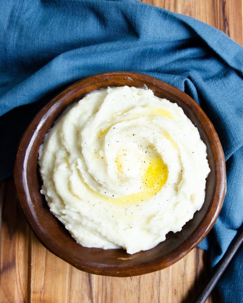 A wooden bowl with mashed potatoes topped with butter and a blue napkin on the side.
