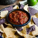Smooth tomato salsa in a small blue dish with blue and yellow tortilla chips around the outside