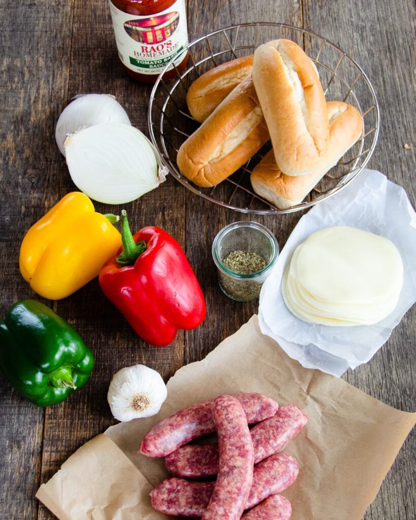 Ingredients for sausage sandwiches on a wooden board - buns, cheese, sausages, peppers, onion and jarred marinara sauce.