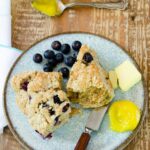 A lemon blueberry scone on a blue plate with blueberries, lemon curd and butter.